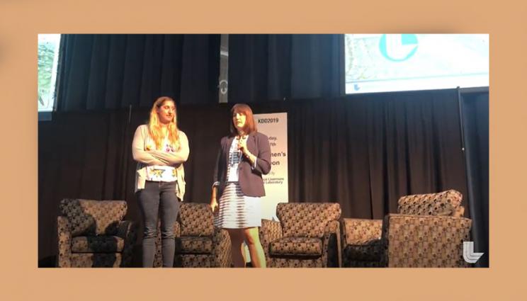 Amanda and Alyson stand on a stage speaking to an audience